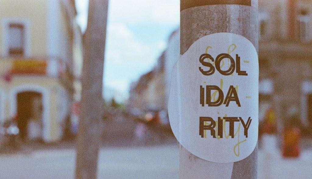 A street photo. In the foreground is a lampost with a circular poster on it which reads "solidarity" in all capital letters.