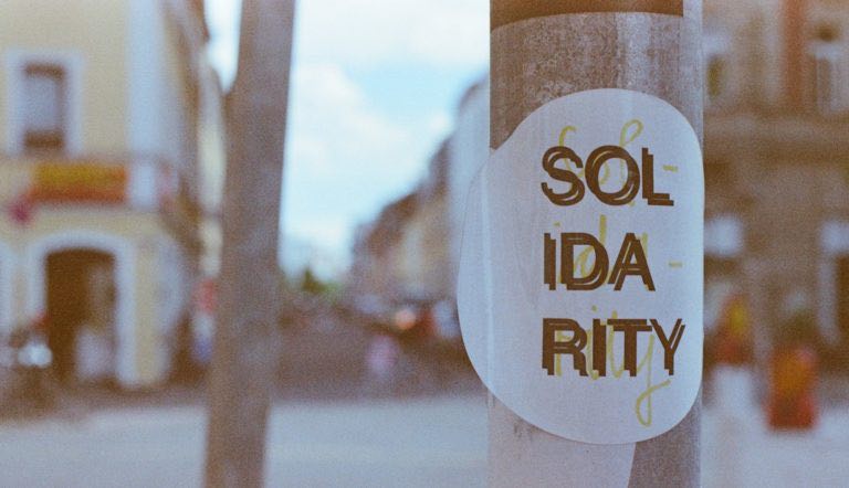 A street photo. In the foreground is a lampost with a circular poster on it which reads "solidarity" in all capital letters.