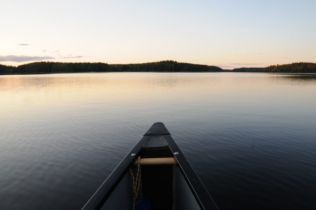 A photo taken from the perspective of inside a canoe. The Canoe is on a large body of water, likely a lake. There is a hint of a sunset, and a black silhouette of land in the distance.