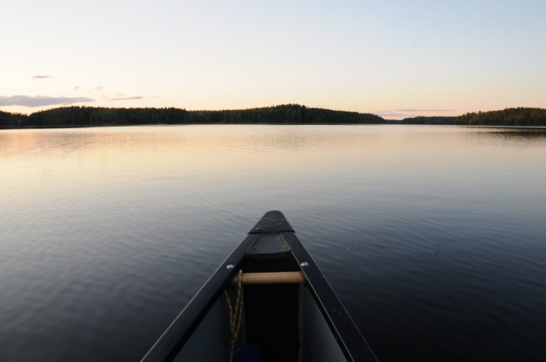 A photo taken from the perspective of inside a canoe. The Canoe is on a large body of water, likely a lake. There is a hint of a sunset, and a black silhouette of land in the distance.