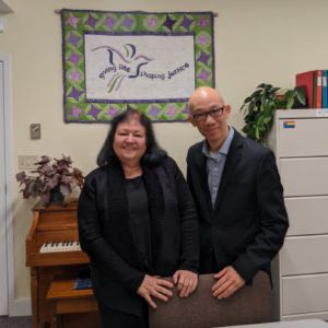 A photo of Alcris Limongi and Alan Lai. Alcris is on the left of the photo, she has shoulder-length black hair and is wearing a dark blue blouse with a black scarf, and is smiling at the camera. Alan is on the right. He is also smiling, and wears glasses, a button-up shirt and a cardigan. In the background is a banner with a dove on it, which reads "giving life... shaping justice." There is also a piano with flowers on top of it and a filing cabinet with a rainbow Pride flag sticker on it and plants atop it.