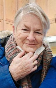 A photo of Pat Stewart. She has short, white hair. She is pulling a white face mask down from her face and smiling warmly. She is wearing a blue parka and knitted scarf.