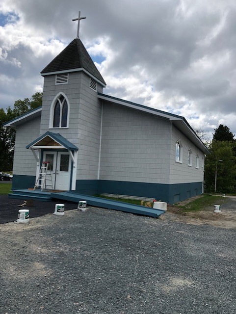 A photo of the church of St. Mary's Sioux lookout. The church is small and beige with a grey roof. There is a steeple at it's front, with a cross atop it
