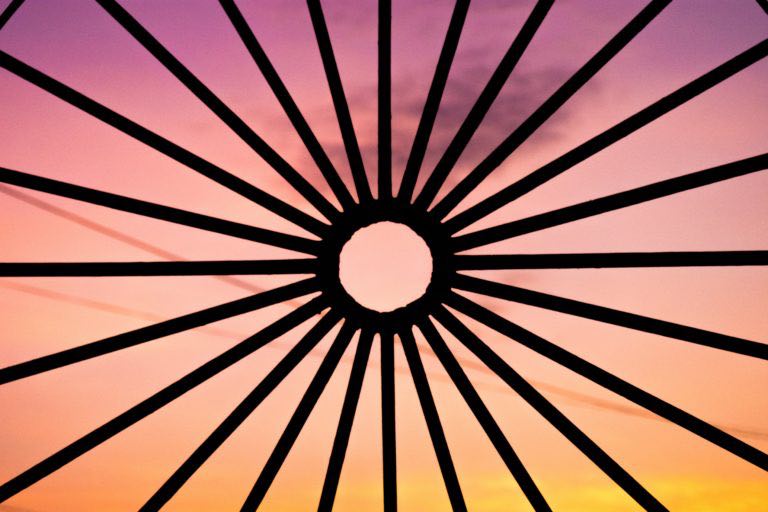 sunset seen through iron bars in the foreground all pointing in towards a circle in the centre made of iron, so that the iron structure silhouette looks like a sun against the backdrop of the sunset