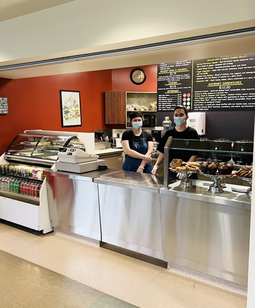 A photo showing Ivan Safarov and Kateryna “Kate” Kharkivska in a cafeteria, behind a counter. They are both wearing face masks and wearing black uniforms, serving gloves, and are looking towards the camera, smiling. The cafeteria has a clean, modern aesthetic. The walls are painted a bright red and an abstract art piece is visible. There is a chalkboard styled menu behind the workers heads. Displays of baked goods and refrigerated goods are visible on the right and left of the photo.