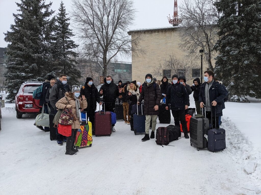 A photo featuring approximately 20 people outside in the snow on St John's College residence in full winter gear and face masks. Each person has various forms of luggage. Some are looking at the camera and smiling, others are looking elsewhere