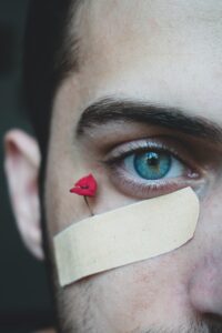 A close-up photo of half of a young, masculine person's face. The person has dark brown hair, and bright blue eyes, one of which is looking into the camera. Beneath the eye is a band-aid with a very small red rose peaking out the top of it.