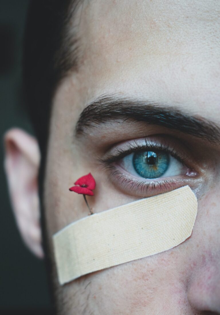 A close-up photo of half of a young, masculine person's face. The person has dark brown hair, and bright blue eyes, one of which is looking into the camera. Beneath the eye is a band-aid with a very small red rose peaking out the top of it.