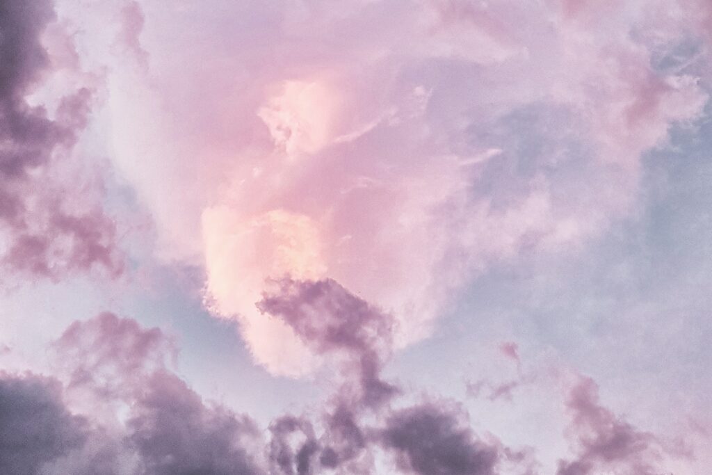 Photograph of clouds in a bright sky. The clouds are shades of lavender and pink against a blue sky.