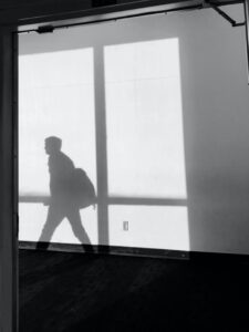 Photo showing a hallway at an angle with bright light coming in through a window against a white wall and outlining the shadow of a young adult, who appears to be walking, wearing a backpack.