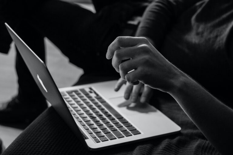 Black and white photo of a person sitting and using a Mac laptop which is perched on their lap.