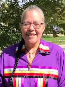A photo of Elder Amanda Wallin set outside. She is wearing a purple, collared top with prominent bands of ribbon ordered in colour: magenta, white, yellow, red, and black. She is also wearing a necklace featuring a shell. Wallin is smiling. She wears glasses. Her hair is black and grey and braided into a long braid which falls over her right shoulder.