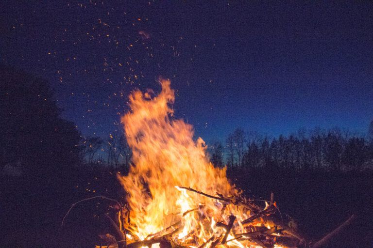 A nighttime photo featuring a deep blue sky and a prominent, large bonfire in the foreground.