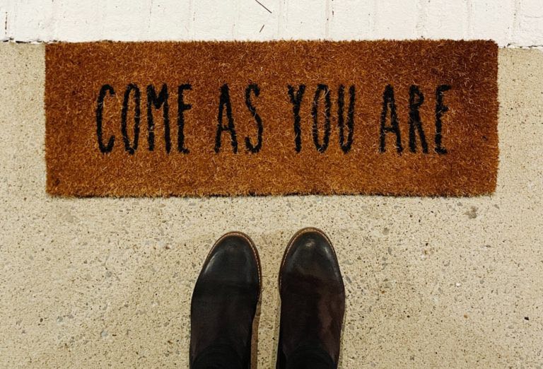 A photo from the perspective of someone looking down at their black boots before a yellow-orange welcome matt with the words "Come As You Are" written in capital letters across it.